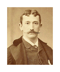 A sepia-toned photograph of a young man with a mustache. He is a white man with curly dark hair parted in the middle, and he has a long pointed mustache. He wears a dark suit and looks directly into the camera.