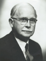 A black and white photograph of an older white man wearing eyeglasses. He is wearing a dark suit and tie, and he has combed-over white hair. He is looking slightly past the camera.