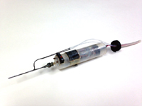 A small surgical tool made of plastic and metal. The tool is shaped like a large plastic syringe with a long metal needle sticking out of one end, and black mechanical parts can be seen through the clear syringe body.