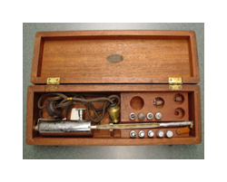 A metal medical instrument sits in a hinged, wooden case. The tool has a silver-colored handle and a long, thin cylinder with a silver metal end. The box also contains several other silver metal ends and a brown power cord.