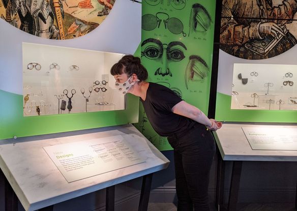A woman looks at a museum display of eyeglasses. She is leaning over a white table towards a plastic case. The gallery has green walls with historical images of eyeglasses printed on them. She is a young white woman with her dark hair pulled into a bun on the top of her head. She is wearing dark clothes and a face mask with a print of eyeballs on it.