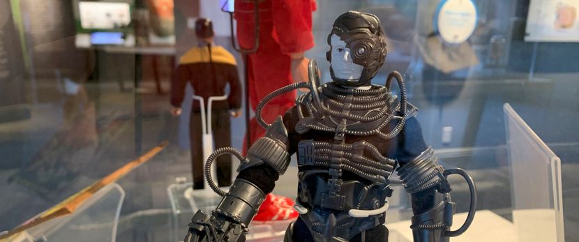 An action figure of a mechanical man from the show Star Trek. His face is white and he wears gray helmet over his head. The helmet also covers his right eye with a small laser. He is wearing a black outfit covered in gray tubes.