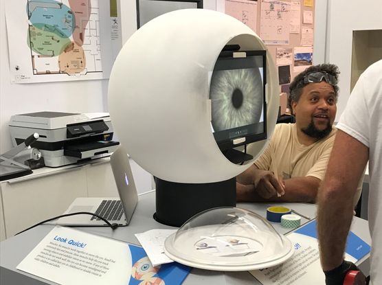 A man assembles a large model of the human eye. He is a Black man wearing safety goggles, and he is installing a TV screen with an image of a human pupil on it into a large white plastic sphere. A small clear plastic dome sits in front of the model.