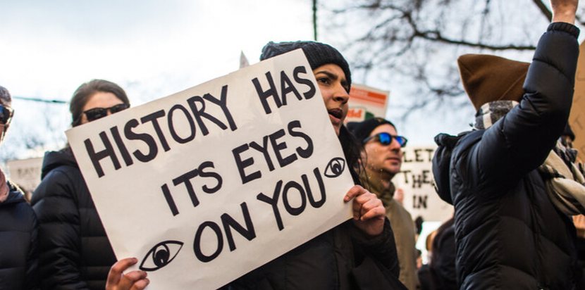 A woman holds a paper sign at a protest. She is in a crowd of other people, and she has dark skin and wears a black beanie. She is holding a white paper sign with black hand-drawn text next to her face. The sign has two drawings of human eyes and reads: History Has Its Eyes on You.