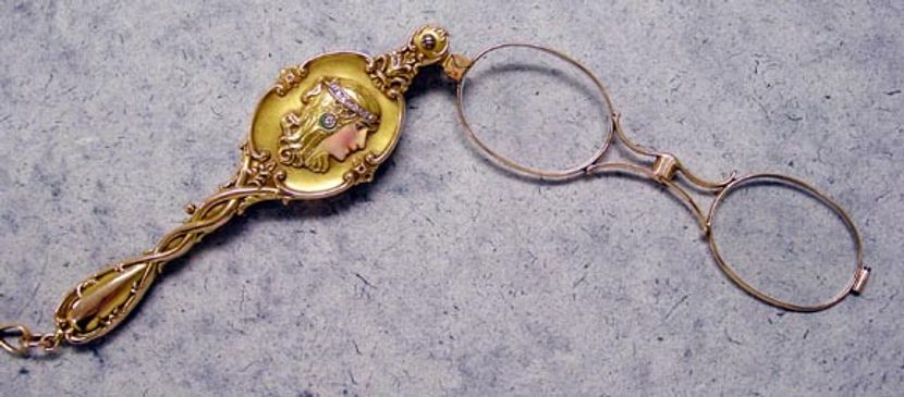 A pair of gold eyeglass lenses attached to a gold handle sits on a gray background. The handle has a decorative border and an image of a woman's face in profile. There is a small diamond inlaid in the woman's hair. The lenses and handle are connected by a hinge.