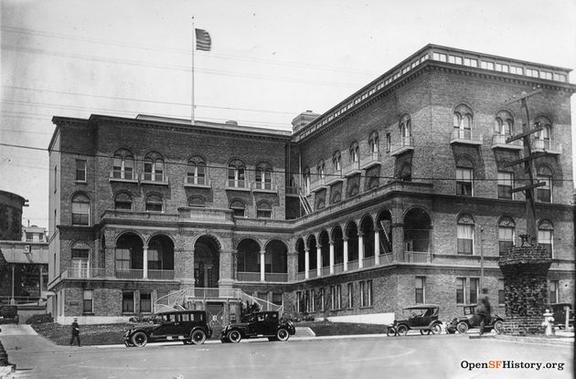 A black and white historical photo of a brick hospital building. The building has three stories with arches and columns in the second floor. It also has a blurry, waving American flag mounted on the top floor. In front of the building, there are four old Model T style black cars parked along the street. Two blurry men walk in front of the cars. In the bottom right hand corner, there is superimposed text that reads OpenSFHistory.com