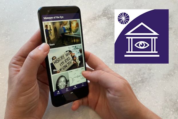 Two hands hold a cellphone displaying the Museum of the Eye app. The hands have light skin, and the phone is black against a gray background.  The app screen reads Museum of the Eye and has several color photographs of people. A purple and white logo is superimposed in the upper righthand corner. it is shaped like a house with columns and has the outline of a human eye in between the columns.