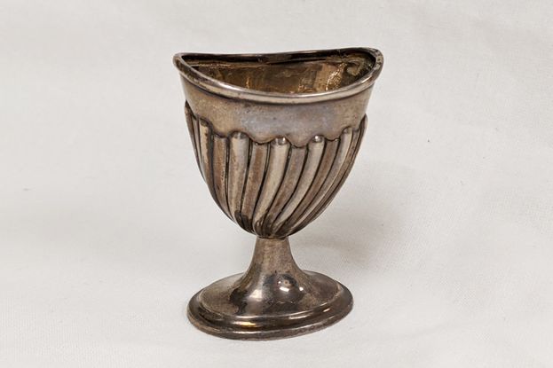 A small metal cup sits on white background. The cup is silver-colored, is a few inches tall, and is made up of a small oval-mounted cup on a round base. The cup portion has a fluted design of vertical lines around it.