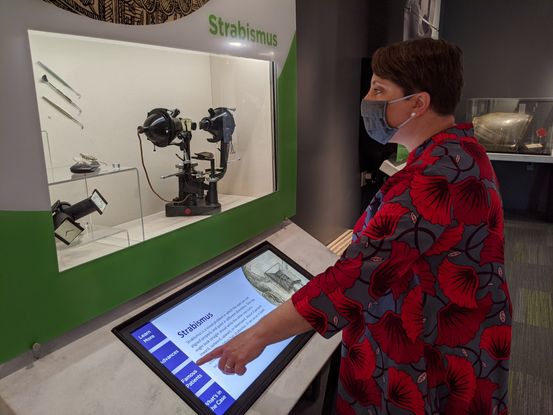 A woman touches a touchscreen while looking at an artifact display. She is a white woman with short cropped brown hair wearing a red and blue floral coat and a face mask. She is touching a touchscreen with her finger while looking at a large black metal medical divide in a clear plastic case. The sign over the case reads: Strabismus.