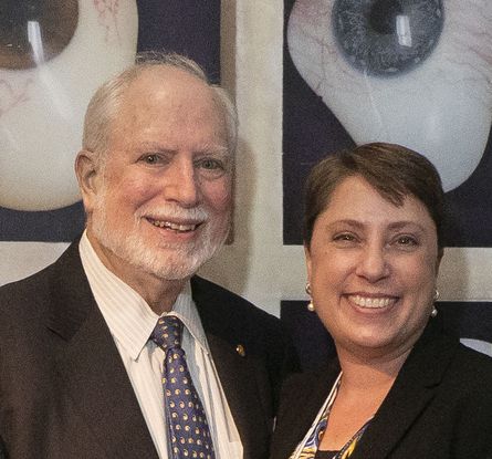 A man and a woman smile in front of a mural of artificial eyeballs. He is an older white man with white hair and a white beard, and she is a white woman with short cropped brown hair wearing pearl earrings. They smile in front of an image of two plastic eyeballs, one with a brown iris and one with a blue iris.