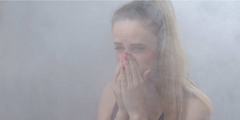 A woman covers her mouth in a smoky room. She is a white woman with hair in a ponytail and is wearing red nail polish. She is surrounded by white smoke, and she is squinting and covering her mouth with her hands.
