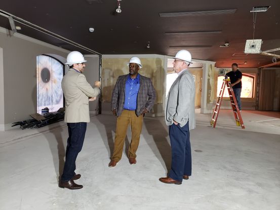 Three men wearing white plastic hardhats stand in a room under construction. Two of the men are white and wearing beige blazers, and the man in the middle is Black and wearing a blue shirt and a checkered blazer. In the background, there is a window covered with an image of a blue eye. There is also a man standing on a red ladder behind the other men.