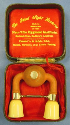 An orange rubber false medical device sits in a red hinged silk case. The device is made up of two beige eye cups connected by an orange rubber hose with a squeeze bulb in the middle. The yellow text in the top of the case reads The Ideal Sight Restorer in cursive script.