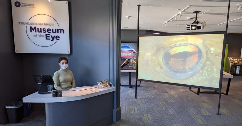 A volunteer sits at the museum front desk. The volunteer is a young white woman with close cropped brown hair, and she wears a green turtleneck sweater. The desk is white and great, and a large white sign hangs above her that reads Truhlsen-Marmor Museum of the Eye in purple lettering. Past her, there is a freestanding video screen with an image of a frog's eye projected onto it.