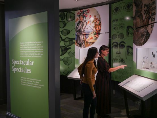 Two women look at a museum display of eyeglasses. The museum gallery has green walls and the green wall behind the women reads Spectacular Spectacles in white letters. The other walls have historical images of eyeglasses printed on them. One woman wears a yellow sweater and has long dark hair. The other woman wears her dark hair in two braids. She is pointing towards a lit case of eyeglasses.