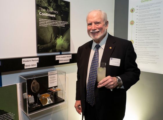 A nicely dressed older man stands and smiles near a museum display. He is a white man with white hair and a white beard. He is wearing a black suit and a blue tie, and he holds a drink with a lime in his hand. He stands next to an artifact case and a sign that reads: The Journey Continues