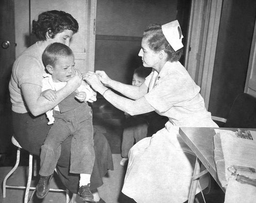 A black and white photograph of a nurse administering a vaccine to a crying child. The nurse is a woman dressed in a white dress and cap, and she administers the shot to a little boy wearing overalls. He is being held by his mother, who is a dark haired woman in a skirt and sweater with a short 1960s hairstyle.