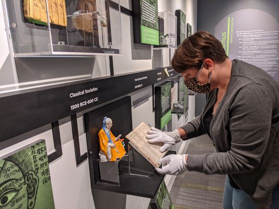 A museum worker installs a book in a display case. She is a white woman with short brown hair wearing a gray cardigan and white cotton gloves. She is placing a small book in a stand next to a ceramic statue of a man. The case is along a wooden timeline on the wall that reads: Classical Societies 1500 BCE - 600CE.