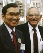 A older photograph of two men in suits. The man on the right is an older white man with white hair and eyeglasses. The man on the left is a middle aged Japanese-American man with black hair and eyeglasses. They both smile at the camera.