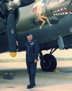 An older man in a blue military uniform stands in front of a military airplane. Only part of the body of the plane and two propellers are visible, but the plane is green with a small painting of a woman on the nose. The man is an older white man in a blue Air Force uniform.