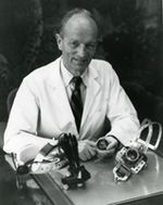 A black and white photograph of a man in a lab coat sitting at a table of medical instruments. He is an older white man with a high forehead, and he is wearing a white lab coat and a black tie. He is sitting at a table with several large black metal instruments on it and smiling at the camera.