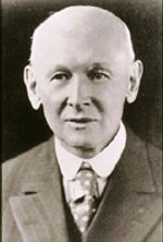 A black and white headshot of an older white man. He is wearing a suit with a spotted tie, and is either bald or has light white hair. He is looking slightly to the left of the camera.
