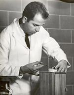 A black and white photograph of a man in a lab coat using a small medical tool. He is a middle aged white man with dark hair and long sideburns, and he wears a white labcoat and a dark tie. He is using either a small black syringe or a small black pair of surgical scissors.