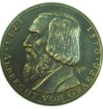 A metal coin with an image of man in profile. The man has longer, collar-length hair and a long beard and mustache. The text across the bottom edge of the coin reads: 1828 Albrecht von Graefe 1870.