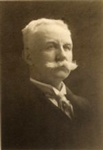 A sepia-toned photograph of a man with a large white mustache. He is wearing a dark suit and an ascot-style tie. He is an older man with white hair and a large white handlebar mustache.