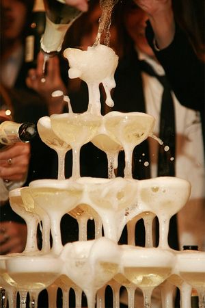 Many clear, glass champagne glasses are stacked on top of one another resembling a pyramid. In the background, bodies wearing black suits pour a golden, bubbly liquid from glass bottles. The liquid is filling each champagne glass and pouring down into the bottom layers of the pyramid. Bubbles fly off the pyramid festively. 
