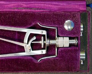 A silver metal medical device sits in a purple velvet case. The device has a small metal plunger attached to two handles.