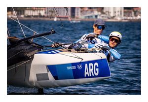 A photograph of two athletes sailing a catamaran sailboat. The sailors are both white and wearing blue uniforms with the letters ARG on them. One athlete wears sunglasses and a baseball cap, and the other wears sunglasses and a helmet. The boat has a white body with blue portions that read: Tokyo 2020 ARG. The athletes are holding ropes and and leaning out off of the boat over the blue water. 