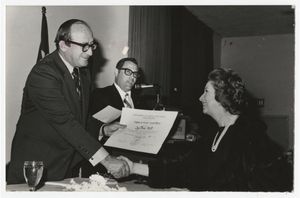 A black and white photograph of two men presenting a woman with an award. The woman is seated, and she wears a black dress and large string of white pearls. She is shaking the hand of a middle aged man with dark hair and eyeglasses, and he is handing her a large sheet of paper. Another dark headed man with eyeglasses smiles on from the background.