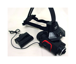 A black plastic headset with a large telescope lens on the front. The headset has an elastic strap meant to go over the head to hold the telescope against the eye. There is a black power cord coming from the telescope lens.