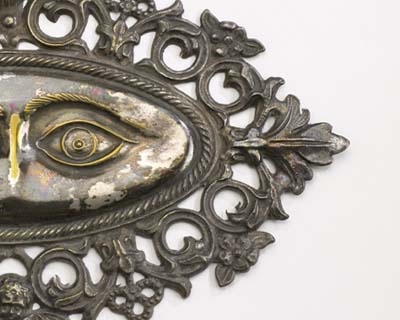 Half of a metal plaque with an image of a human eye stamped on it. The metal is silver-colored with some tarnish. The human eye is inside of an oval, and there is a floral decorative border.