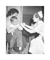 A black and white photograph of a nurse administering a shot to a child. The nurse is a young, white woman wearing a white dress and white cap. She is giving a shot in the arm of a young, crying child wearing overalls. The child is being held by a woman wearing a sweater and a skirt, with short dark hair in a 1950s bob hairstyle.