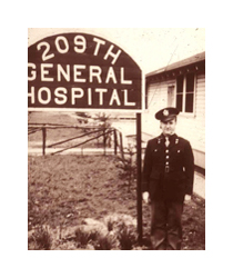 A black and white photograph of a man in military uniform standing between a white wood building and a large wooden sign. He is a young white man wearing a dark uniform with his uniform hat cocked to the side. The large white lettering on the sign reads: 209th General Hospital.