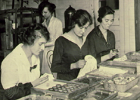 A black and white photograph of three woman cleaning small medical objects. All of the women have dark hair pulled up in 1930s hairstyles, and two of them wear black dresses while one woman is dressed in white. They are cleaning small glass objects from wooden boxes on the table in front of them.