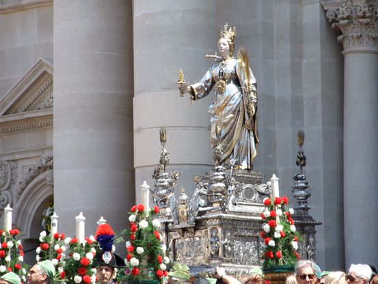 A statue of a woman wearing silver metal robes on a large silver platform being carried on the shoulders of men. The statue wears a crown and holds a palm frond in one hand and a flame in the other. The platform is lined with candles and red and white flowers. 