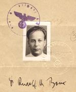 A page of a passport with a small photograph, a signature, and a seal. The photography is of a white middle aged man with dark hair. His signature is below in ink and reads: Rudolf H. Bock. There is a purple seal stamped over the corner of the picture in the shape of a circle with an eagle holding a Nazi swastika inside.